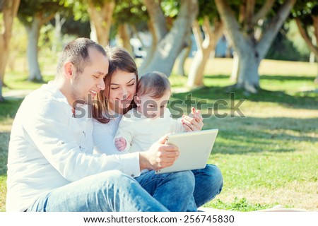 Happy family and digital technology. Young parents and little kid using table computer while sitting on grass in the park.