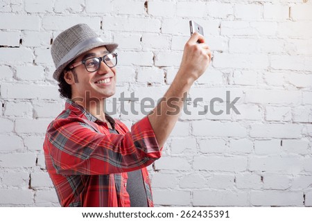 Cheerful selfie. Handsome young Arabic man in shirt holding mobile phone and making photo of himself while standing against brick wall