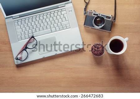 Overhead of modern comfort work place. Different objects on wooden background. Items include camera, glasses, laptop, cup of coffee and muffin