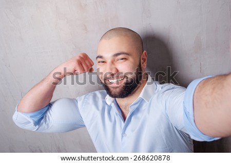 Sport and selfie! Handsome young latino man in shirt holding camera and making selfie and showing his strong muscles while standing against grey wall.