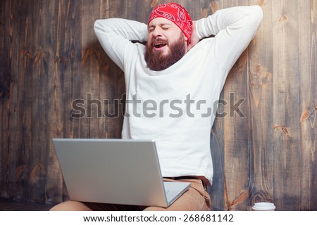 So tired! Young bearded man working on laptop and yawning while sitting against wooden wall.