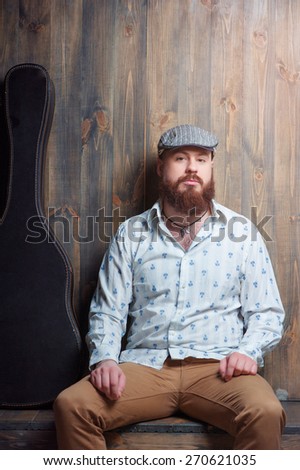 Portrait of musician. Handsome young bearded man sitting with guitar case over wooden background.