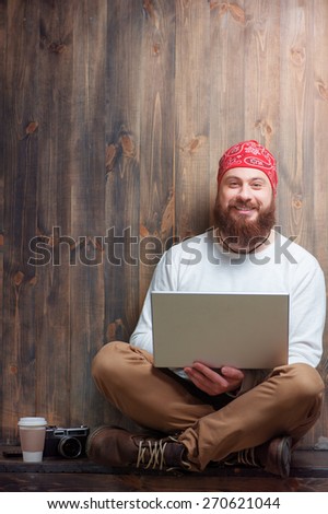Comfort workplace. Smiling bearded man using laptop while sitting over wooden background.