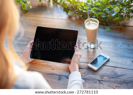 Working at the cafe. Close up of female hands holding tablet computer on the wooden table with glass of latte and smartphone on it.