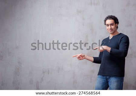 pointing copy space. Happy young Arabic man in casual wear looking at camera and smiling while standing against grey background and pointing away