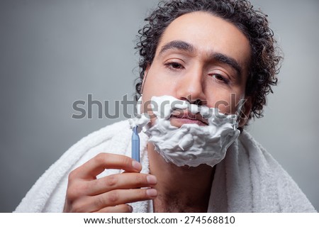 Man shaving. Handsome shirtless Arabic man shaving his face and looking at camera while standing against grey background