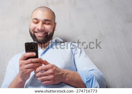 Handsome young man in shirt using mobile phone and smiling while standing against grey wall.
