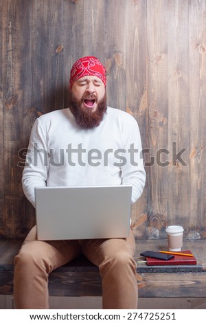 Need coffee for wake up. Yawning bearded man with laptop computer sitting over wooden background.