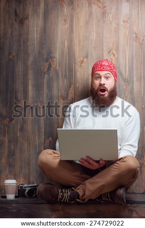 What a surprise! Amazed bearded man using laptop while sitting over wooden background.