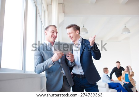 Creative work and technology. Two smiling handsome men using tablet computer while discussing something with their colleagues in the background.