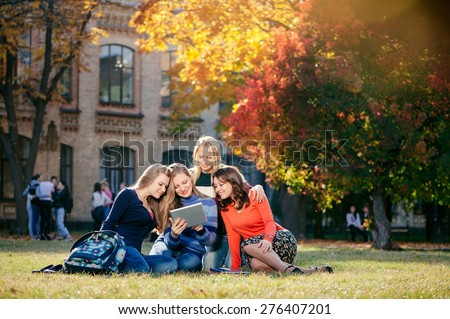 Surfing the net together. Four happy attractive female caucasian students looking at tablet pc and smiling while  sitting on grass at campus together