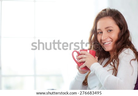 Calm and coziness. Beautiful young woman with cup of tea sitting against the window at home