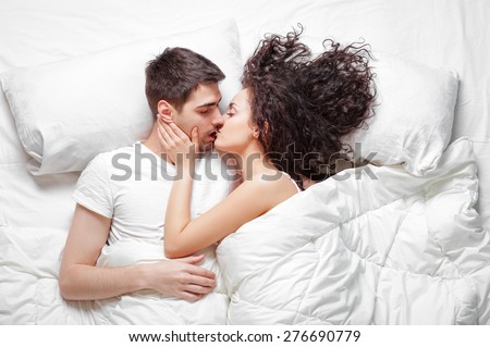 Good morning! Love and comfort. Overhead of happy beautiful young couple lying on the bed kissing and hugging. Top view.
