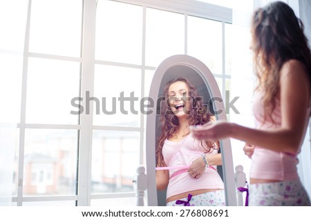 Weight loss. Happy young woman measuring her waist while looking at mirror at home.