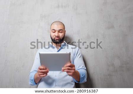Working on tablet computer. Handsome young arabic man working on digital tablet while leaning on grey wall.