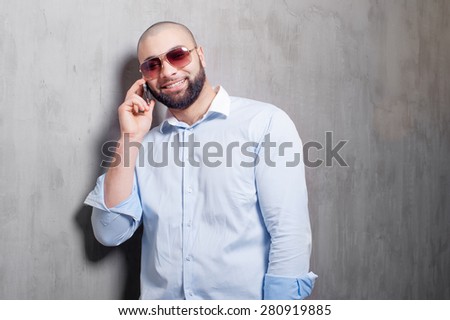 Confident talk. Happy young bearded man talking on the mobile phone and looking at camera while standing near the grey wall.