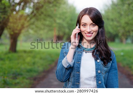 Cheerful conversation. Attractive young woman talking by smartphone while walking by summer park.