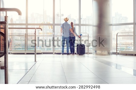 Traveling concept. Back view of loving couple in casual wear standing near the window of international airport terminal.