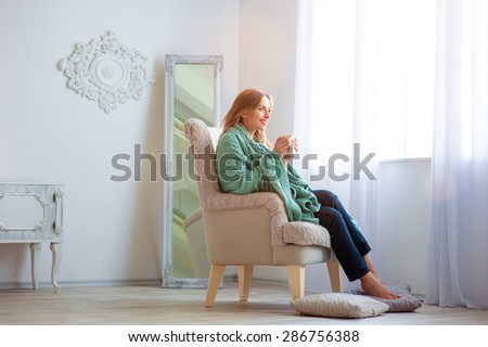 Calm and coziness. Attractive young woman with cup of coffee sitting on armchair at home. Light beautiful interior.