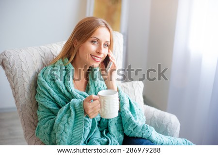 Good talk.Technology and coziness. Beautiful young woman with cup of tea talking by smart phone while sitting on armchair at home.