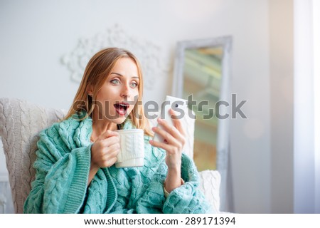 Shocking news.Technology and coziness. Amazed young woman with cup of tea using smart phone while sitting on armchair at home.