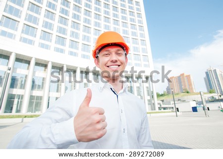 Confident contractor. Cheerful young man in shirt and hardhat showing thumb up and smiling while standing outdoors