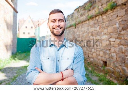Portrait of happy young man in casual shirt keeping arms crossed and smiling while standing outdoors.