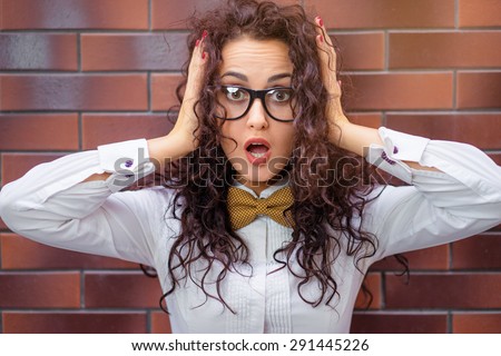 Shock! Amazed young woman with curl hair in glasses holding hands on her head standing against brick wall.