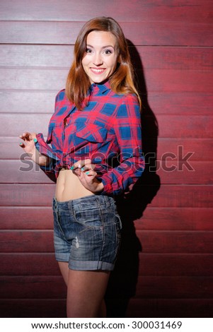 Beautiful young woman in plaid shirt and red glasses showing her belly while standing against the wall.