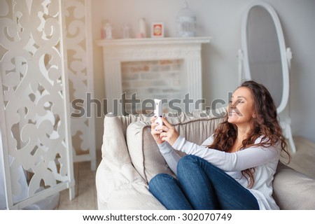 Young attractive woman taking picture with her white smartphone sitting on sofa at home.