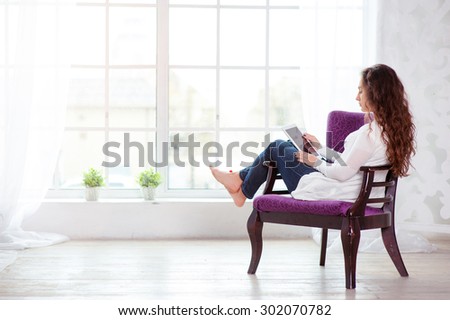 Technology and coziness. Beautiful young woman using tablet computer while sitting at armchair at home.