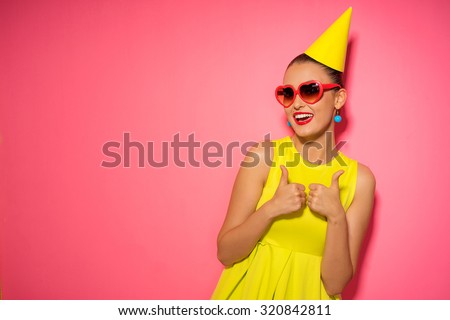Celebration and party. Having fun. Young pretty woman in yellow dress and birthday hat is laughing. Colorful studio portrait with pink background.