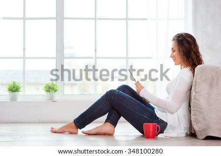 Technology and coziness. Beautiful young woman with cup of tea using tablet computer while sitting on floor at home.