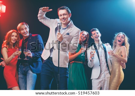 Stand-up comedy and party. Showman with microphone. Group of cheerful friends toasting with wineglasses among confetti.