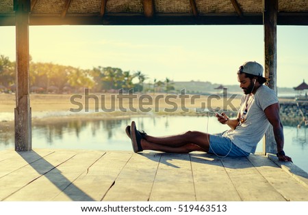 Relaxed and cheerful. Listening to music. Outdoor portrait of happy young african man resting on deck near the sea.