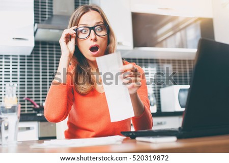 stressed over bills. portrait of surprised young  woman using a laptop computer sitting at her kitchen holding utility bill and bank statements. Home interior.