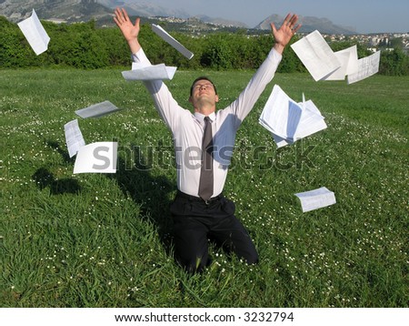 A young businessman is sitting on his knees on a grass and throwing away his papers