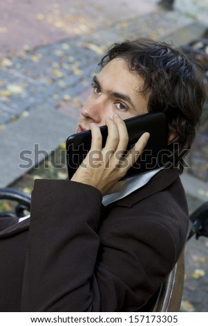 Man in brown suit sitting on the bench and talking on tablet at the park. Young businessman. Talking on the phone. Telecommunications. Gadgets, PC. Business style, a man in business clothes. Brunette.
