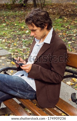 Young man in brown suit sits on the bench at the park and reads digital book
