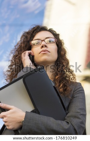 Confident business woman with curly hair with document case talking on phone outdoor