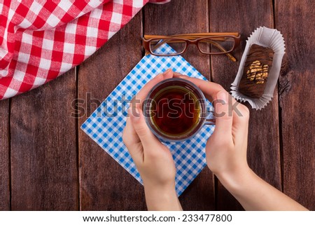 Hands of a girl with a cup of tea. Glass cup of black tea on a wooden table next to a chocolate cake. Glasses, cake, black tea, cloth and hands.