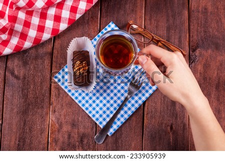 A cup of tea on the table next to the cake and glasses. Chocolate cake with nuts on a table next to a cup of tea and eyeglasses. Female hand touching cup of tea.