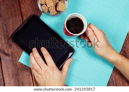 Desk office worker. A young man at work. Tablet PC on the desk. Cup of black coffee, men\'s hands, waffles, and the plate on the table. Top view.