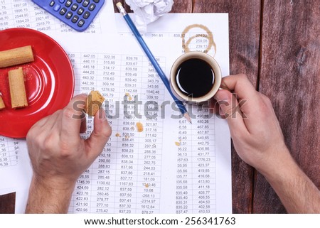 Coffee spots on the paper. Untidy workplace of accountant. Table numbers. Report, debit credit, verification digits. Calculator, wafers, chips, cup of coffee, man\'s hands and documents.