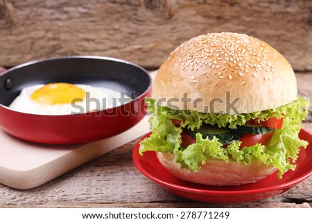Roll with sesame and vegetables. A wooden table. Fried egg on the pan. The Burger on the plate. Sandwich. Lettuce, tomato and cucumber. White fresh bread, and vegetables.
