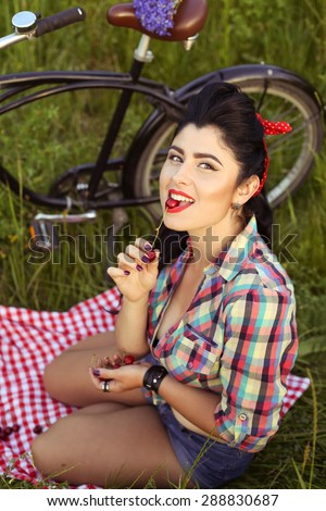 Pin-up girl in a shirt and shorts outdoors. Sexy girl with a Bicycle. Brunette with red lips. Girl eating berries. Sexy, bright, emotional, xxx. Tanned brunette girl with bright makeup. Red lips.