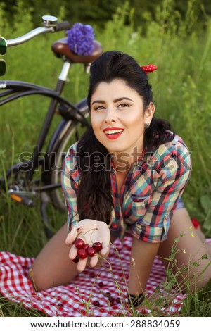 Pin-up girl in a shirt and shorts outdoors. Sexy girl with a Bicycle. Brunette with red lips. Girl eating berries. Sexy, bright, emotional, xxx. Vintage style, fashion look. Retro look. Pin up look.