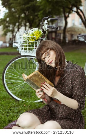 Girl cyclist. Relax on the lawn, sit on the grass. Beautiful young woman in retro dress with a book in his hands. Retro Bicycle with basket of flowers. Retro look. Woman with bike. Street cycling.