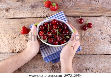 A bowl of ripe cherries and strawberries on the table. A bowl of fruit on the table. Hands with berries. Male hands and red cherries. Cherries, strawberries, bowl, hands, and desks.