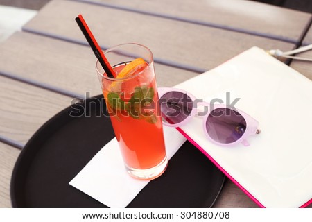 Red cocktail with ice and mint on a tray. Cocktail and sunglasses. Alcoholic cocktail on the table in the lounge area. Lounge cafe. Tinted photo. Tray, cloth,  sunglasses and bag.  Selective focus.